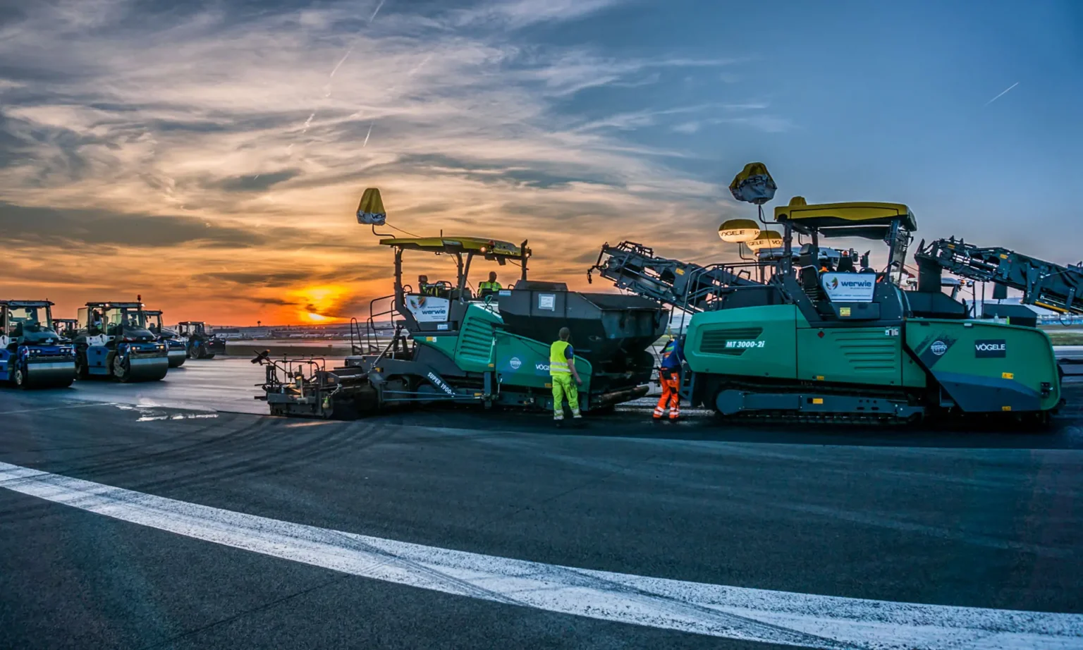 Zwei Vögele Großfertiger der Highway Class beim großflächigen Asphalteinbau auf einer Flughafen-Startbahn bei Dämmerung.