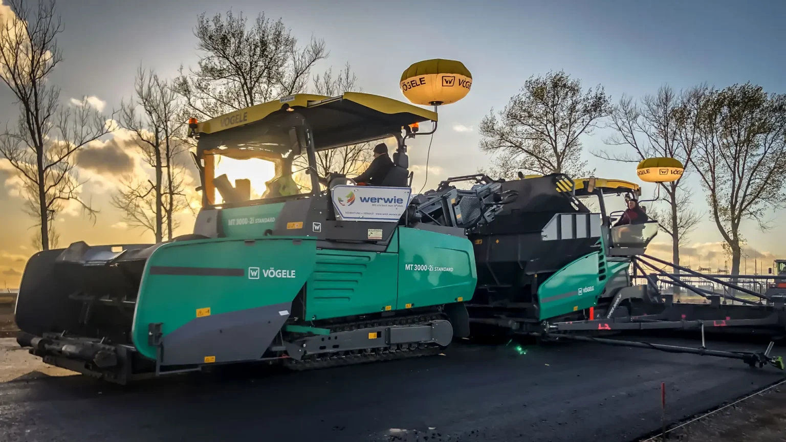 Ein grüner Vögele Standard-Beschicker von werwie im Einsatz bei der berührungslosen Asphaltübergabe an einen Fertiger bei der Dämmerung.